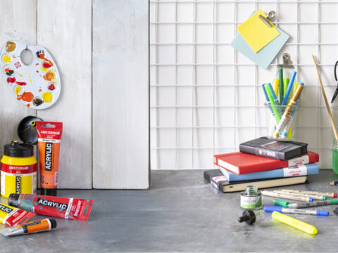 Et bredt utvalg av kunst- og tegneutstyr er plassert på et bord foran lyse veggpaneler, inkludert pensler, malingsrør, akrylfarger, tusjer, skissebøker og fargeblyanter.