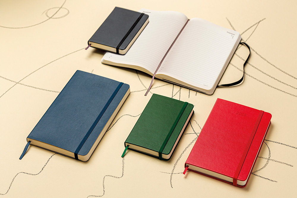 Seks Moleskine-notatbøker i ulike farger og størrelser ligger spredt utover en beige overflate med tegnet mønster.