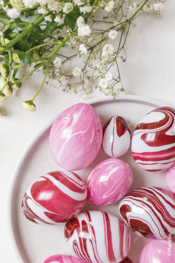 Rosa og røde marmorerte påskeegg på en lys tallerken, med hvite blomsterkvister i bakgrunnen.