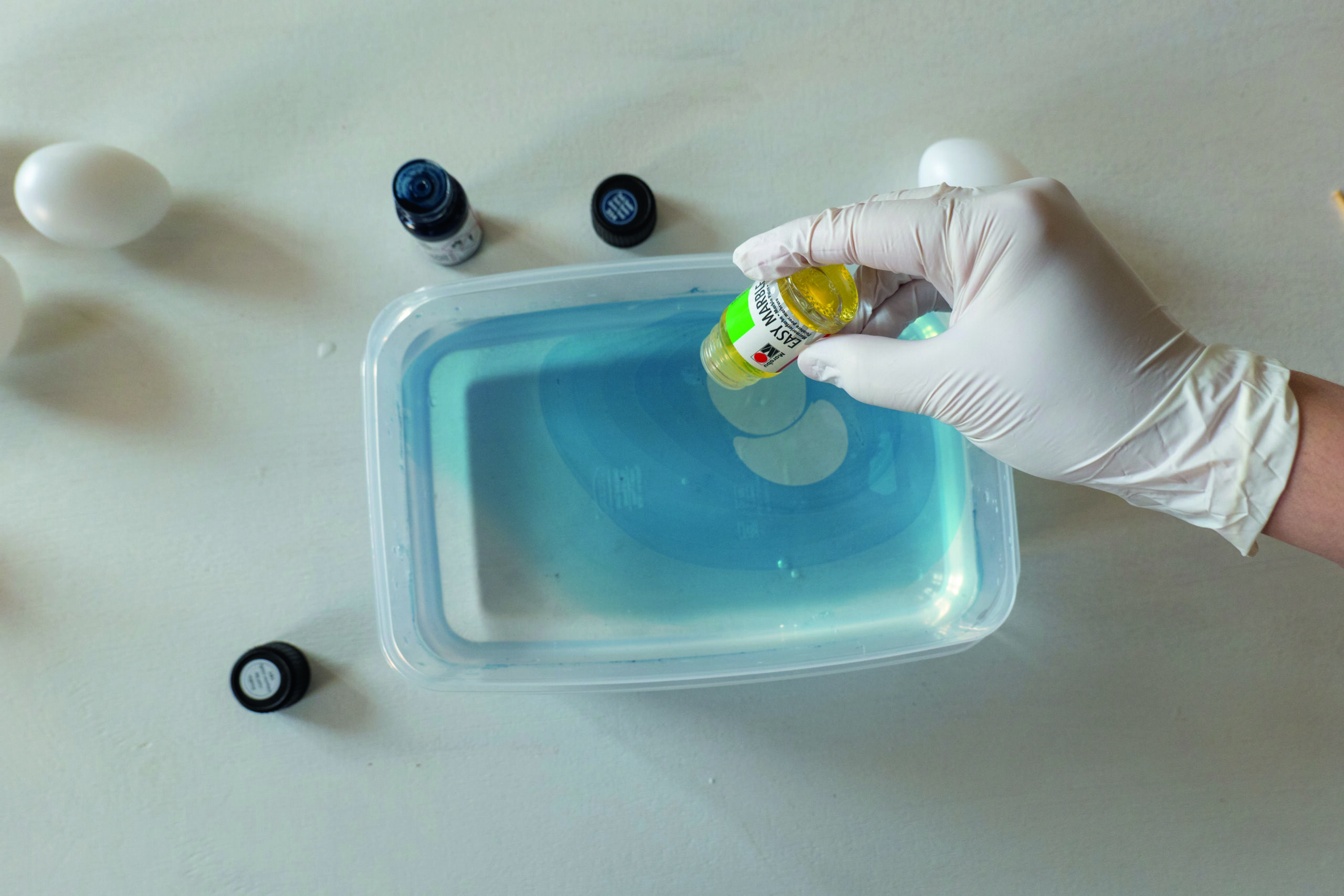 Hånd drypper gul marmoreringsfarge i blåfarget vann i en plastboks.
