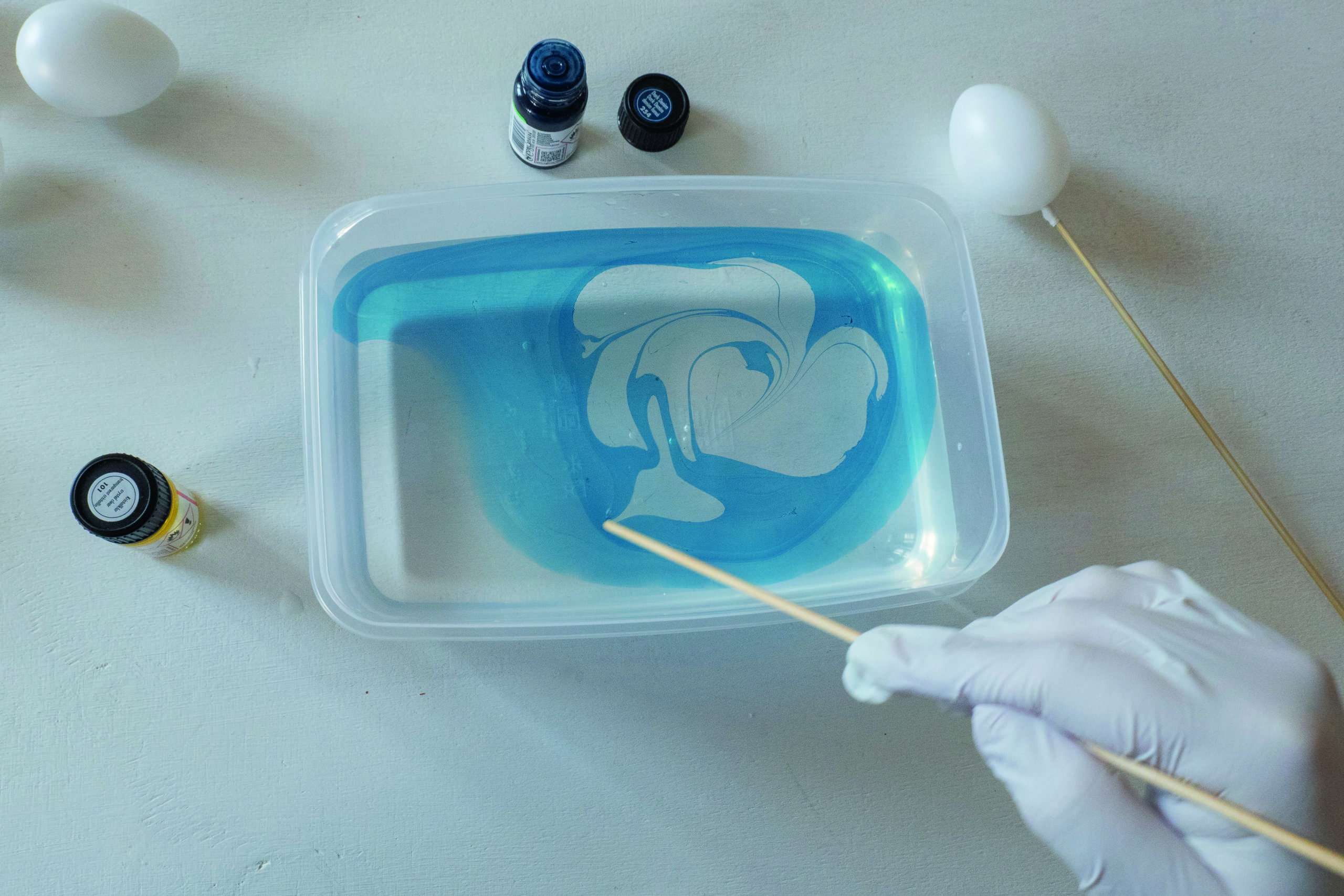 Tannpirker rører blå og hvit marmoreringsfarge i vann i en plastboks.
