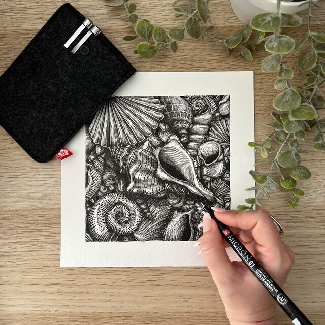 Hånd som tegner detaljerte skjell med sort fineliner på hvitt papir. Rundt ligger en filtmappe, en plante og tegneutstyr på lyst trebord.
