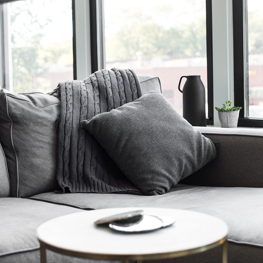 Grå sofa med grått pledd og pute ved et vindu, med et lite rundt bord foran og en plante og kanne i vinduskarmen.