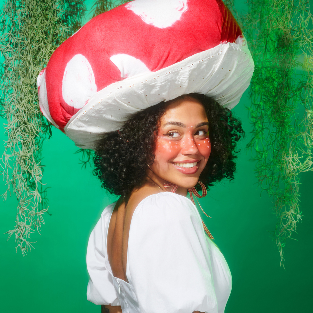 En kvinne kledd i soppkostyme med stor rød og hvit hatt smiler mot kamera foran en grønn bakgrunn med hengende planter.