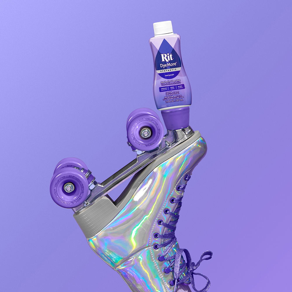 Flaske lilla Rit DyeMore balanserer på en holografisk rulleskøyte med lilla hjul mot ensfarget lilla bakgrunn.