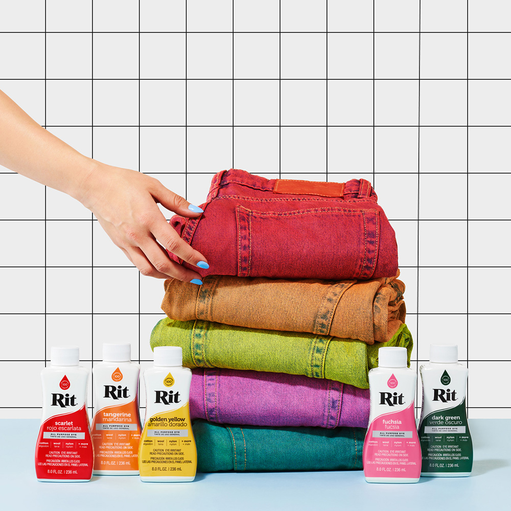 En hånd med blå neglelakk løfter en rød jeans fra en stabel fargerike jeans foran seks flasker tekstilfarge fra merket Rit.