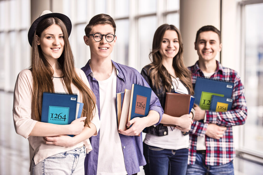 Fire unge studenter står i rekke innendørs med bøker og notatblokker i armene, og smiler mot kameraet.