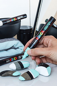 En hånd dekorerer små steiner med en lyseblå Posca-tusj.