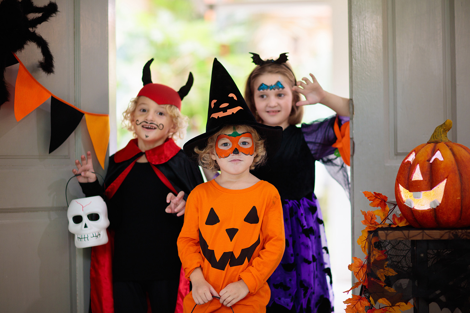 Tre barn i halloweenkostymer med ansiktsmaling, stående i en døråpning ved siden av en gresskarlykt.