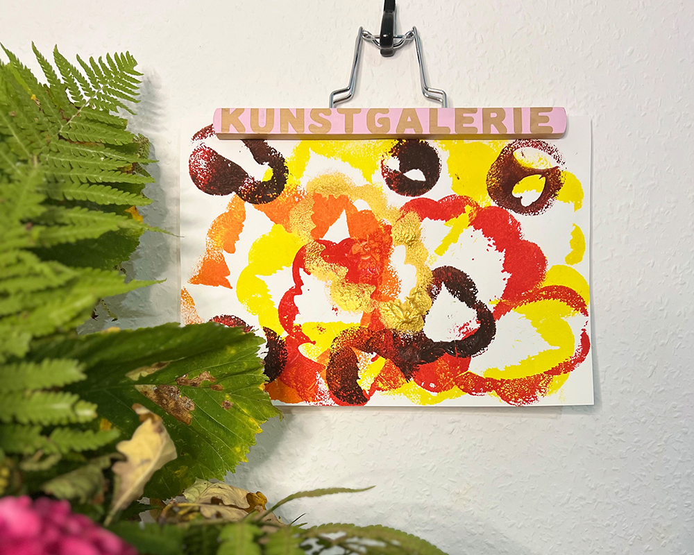 Kunstverk i høstfarger henger på vegg ved siden av blomster og kongle.