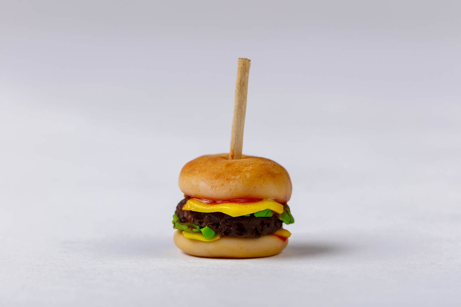 Miniatyrhamburger laget av Cernit med pinne i midten mot lys bakgrunn.