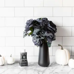 vart vase med mørkfargede blomster på en marmorbenk, omgitt av små hvite gresskar og en flaske tekstilfarge.