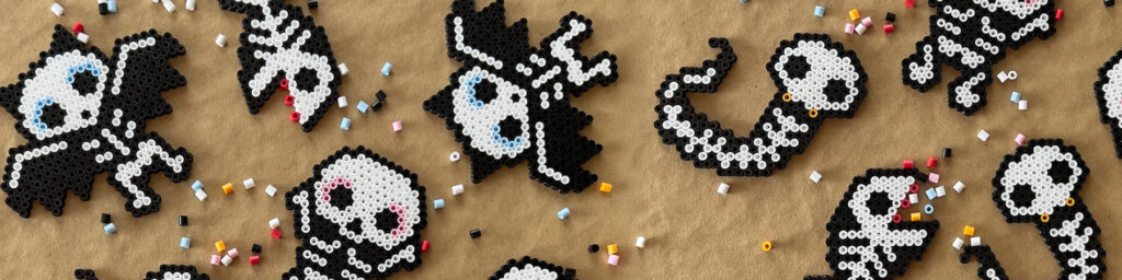 Flere små perlefigurer i svart og hvitt formet som skjeletter, flaggermus og ormer ligger på et brunt underlag med spredte perler rundt.