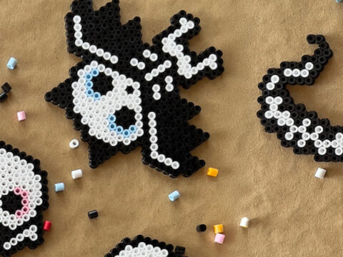 Flere små perlefigurer i svart og hvitt formet som skjeletter, flaggermus og ormer ligger på et brunt underlag med spredte perler rundt.