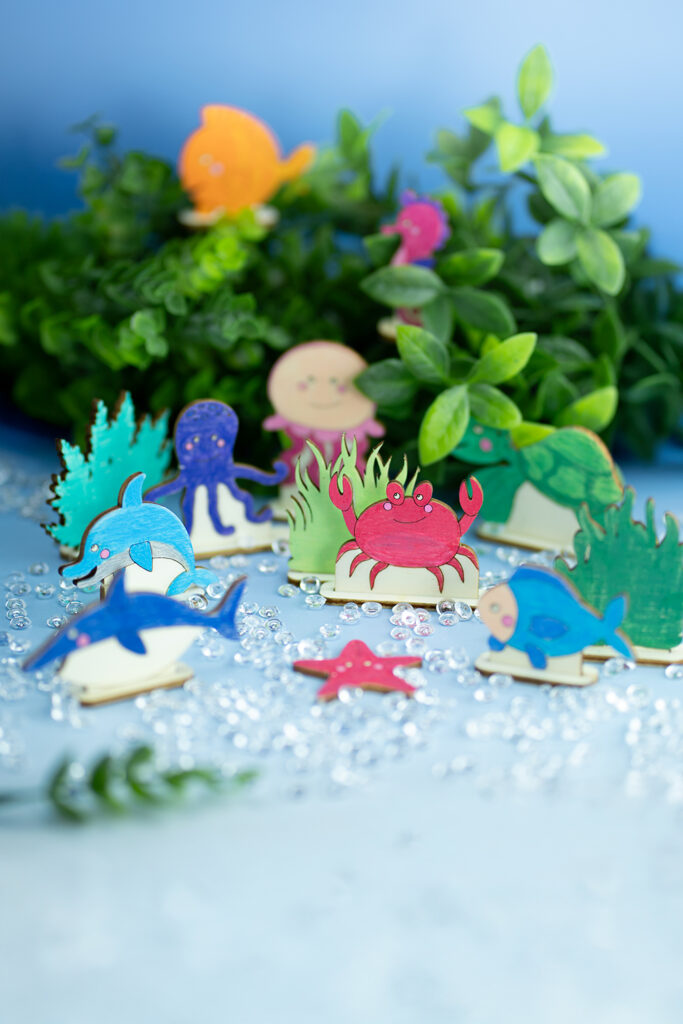Fargerike trefigurer av havdyr står på et blått underlag med grønne planter i bakgrunnen.