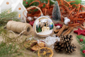 Julekule med vinterlandskap i tre omgitt av julepynt, kongler og kanelstenger.