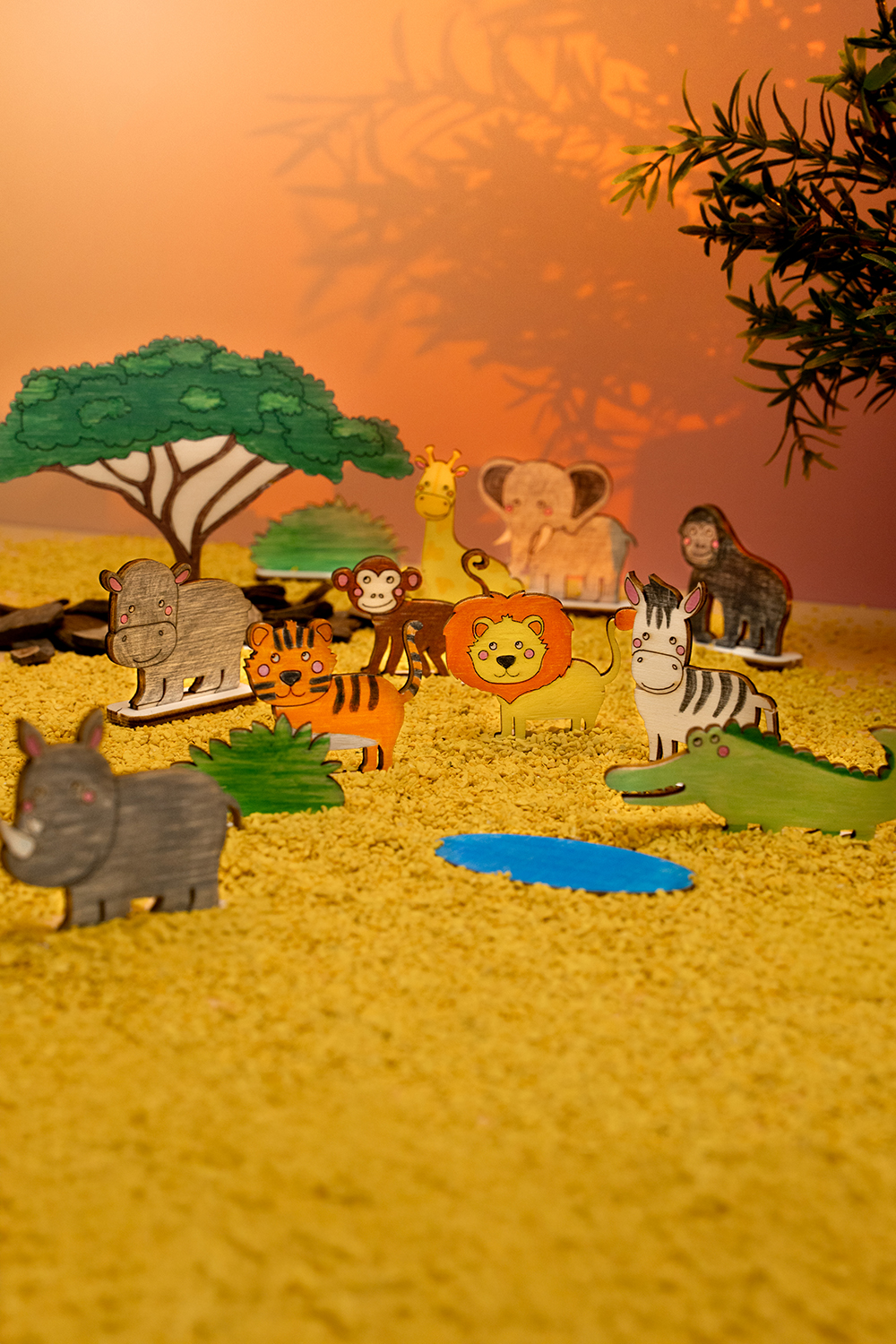Fargerike trefigurer av savannedyr står på gul sand foran oransje bakgrunn.