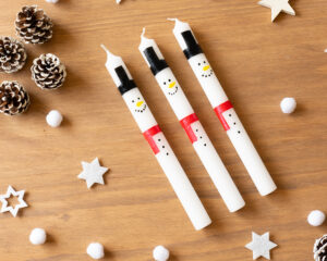 Tre hvite lys dekorert som snømenn med hatt, skjerf og ansikt malt på.