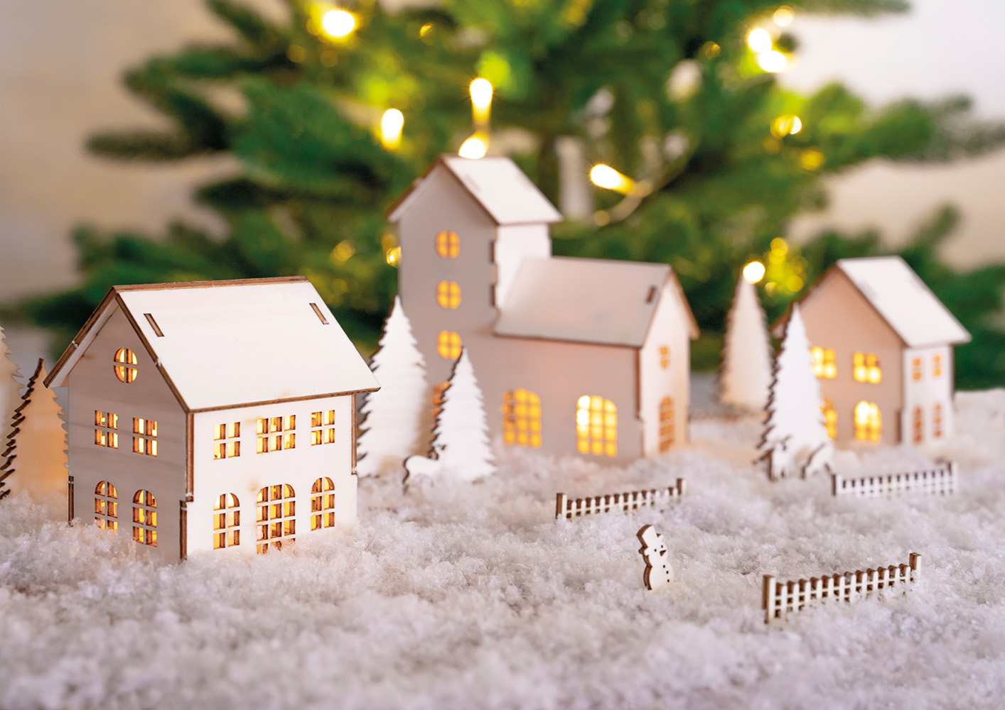 Små trehus med lys står i kunstig snø foran et juletre med varme lys.