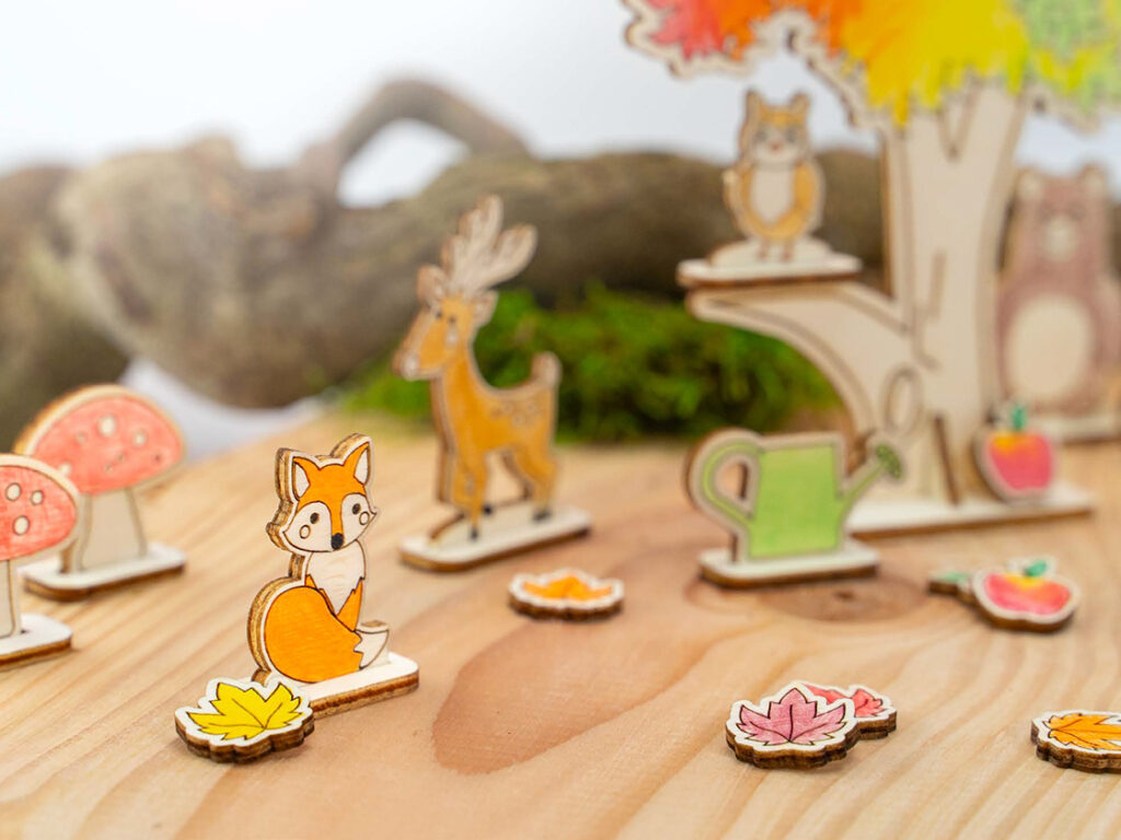 Små, fargerike trefigurer med høstmotiv står på et trebord, inkludert rev, rådyr, blader og sopp.