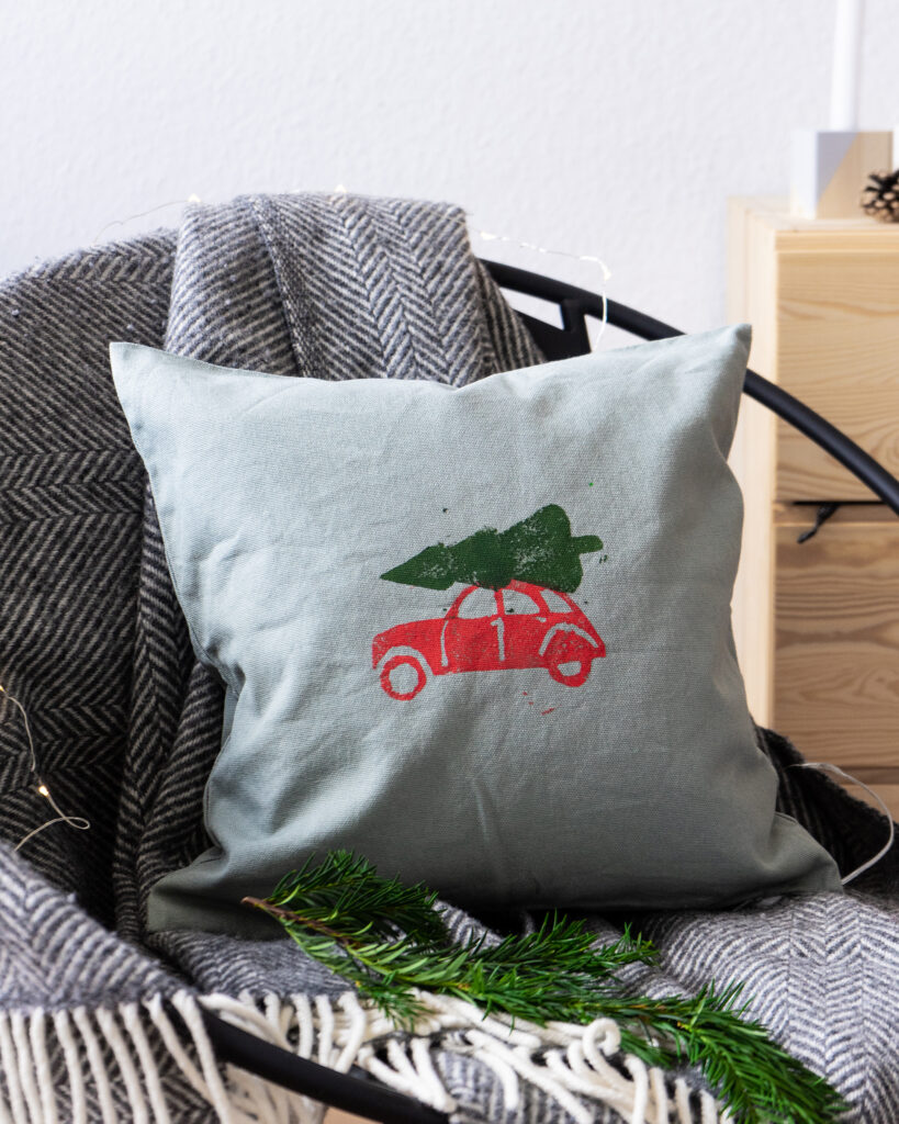 Grønn pute med rødt trykk av bil som frakter et grønt juletre.
