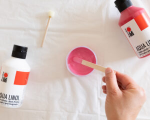 Hånd som rører rosa trykkmaling i en liten beholder med en trepinne.