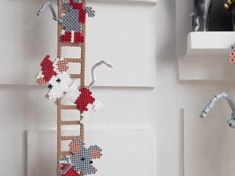 Flere Hama-perlemus i rødt, grått og hvitt klatrer på perlestiger mot lys vegg.