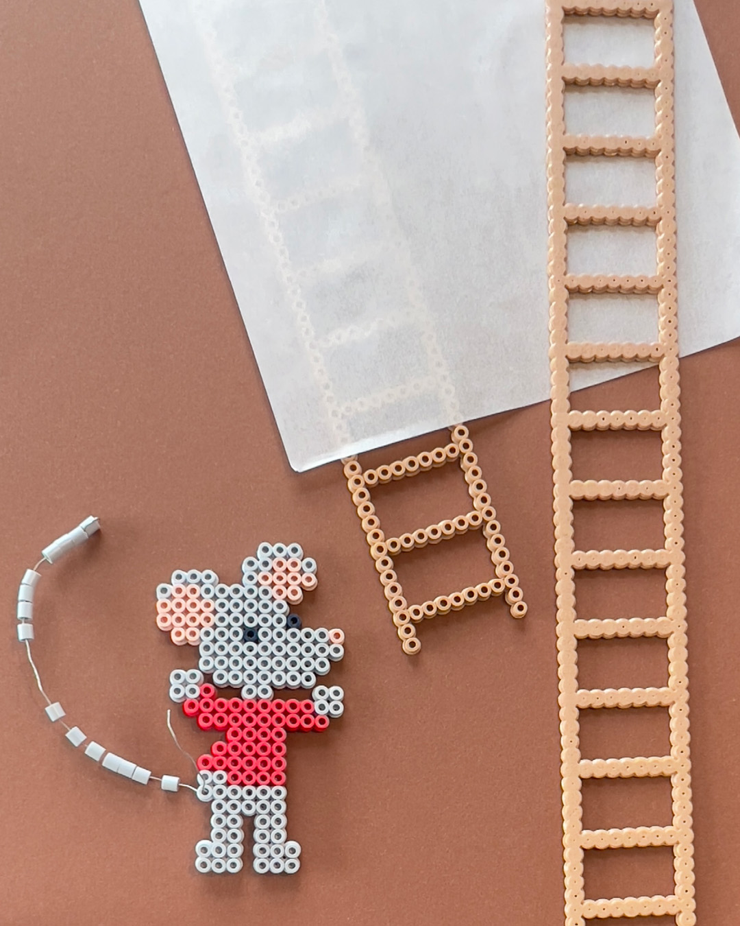 Mus og stiger laget av Hama-perler ligger på en brun flate sammen med bakepapir.
