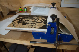 Tresnittmotiv ligger i en blå trykkpresse med papiret delvis løftet.