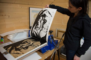 Kvinne løfter et ferskt tresnitt fra en blå trykkpresse i et trepanelt verksted.