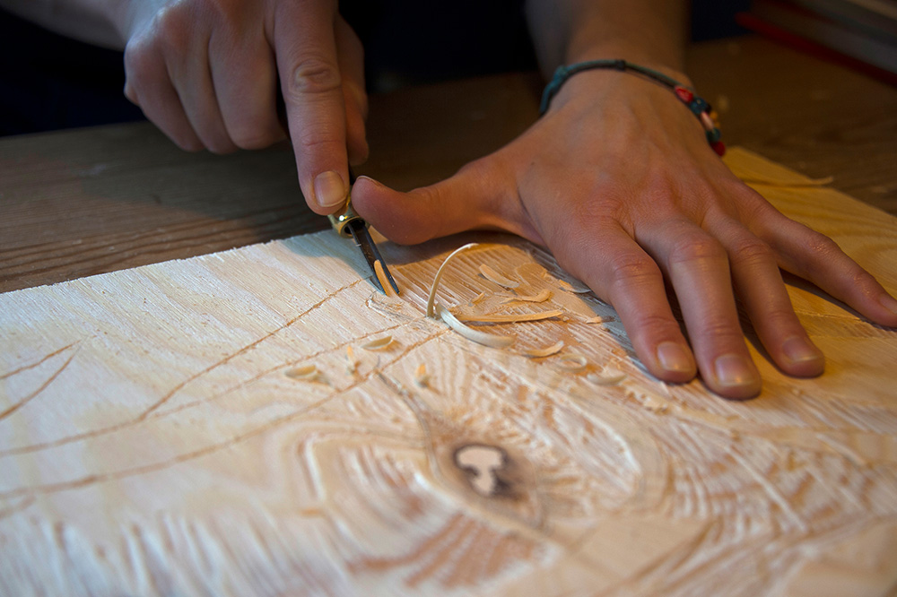 Hender skjærer ut linjer i en tresnittplate med et lite jern.