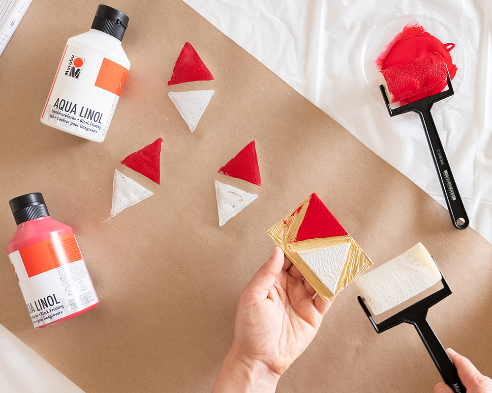 Hender som trykker røde og hvite trekantformer med en utskåret plate på brunpapir.