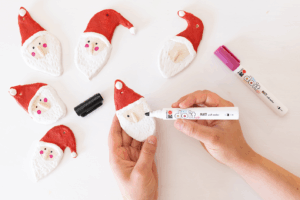 Hender tegner ansikt på nissefigur i trolldeig med tusj, omgitt av flere ferdige figurer.