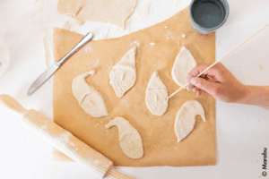 Hånd former detaljer i uferdige nissefigurer av trolldeig på bakepapir, sett ovenfra.