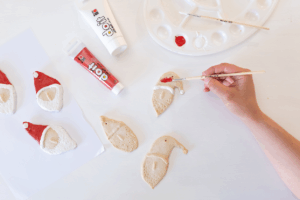 Hånd maler røde detaljer på nissefigur i trolldeig med pensel, sett ovenfra på hvit flate.