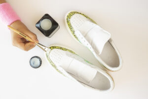 Hånd påfører lim med pensel på hvite sneakers med gult mønster langs kanten.