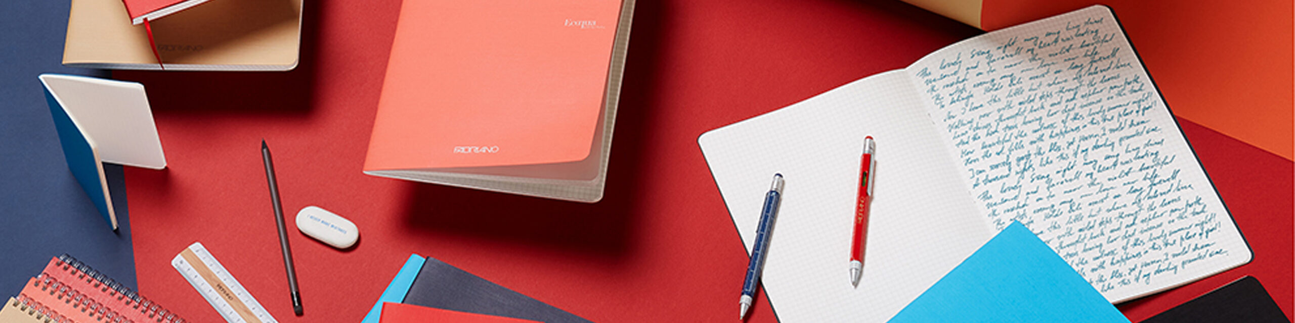 Oppslått notatbok med håndskrevet tekst, penner og skrivesaker arrangert på rød flate.