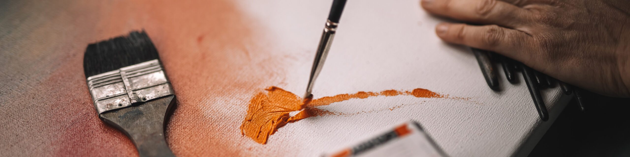 Nærbilde av pensel som påfører oransje maling på lerret, med bred pensel ved siden av.