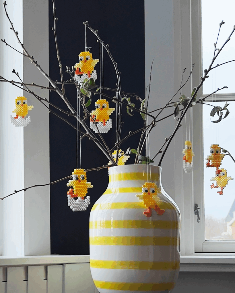 Gule kyllinger av Hama strykeperler henger på greiner i en gulstripet vase i et vindu.