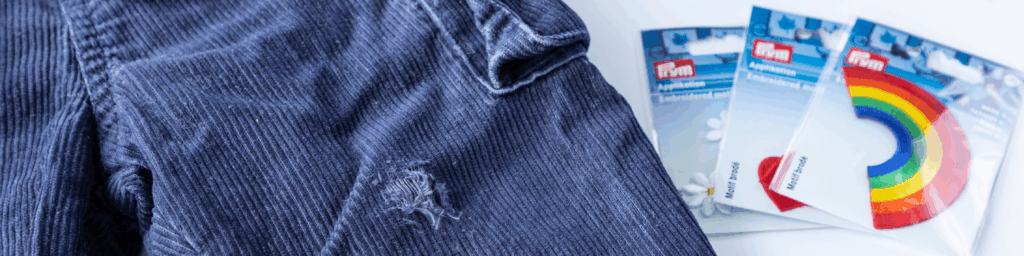 Blå kordbukse med hull ved siden av Prym reparasjonslapper i emballasje.