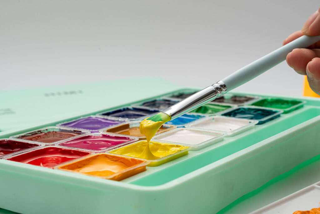 Hånd holder pensel med gul gouache over Himi malepalett med flere farger