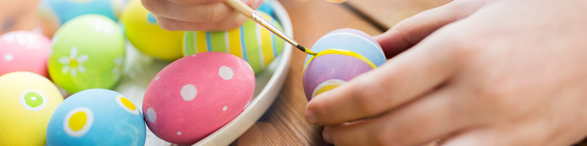 Hånd maler dekor på fargerikt påskeegg med pensel, flere malte egg ligger i en skål på bord.