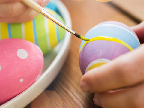 Hånd maler dekor på fargerikt påskeegg med pensel, flere malte egg ligger i en skål på bord.