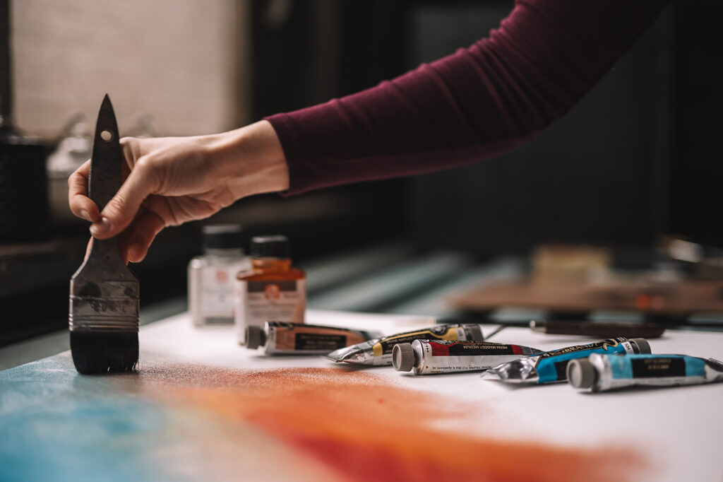 Hånd holder bred pensel og maler på papir med farget pigment, malingstuber ligger på bordet
