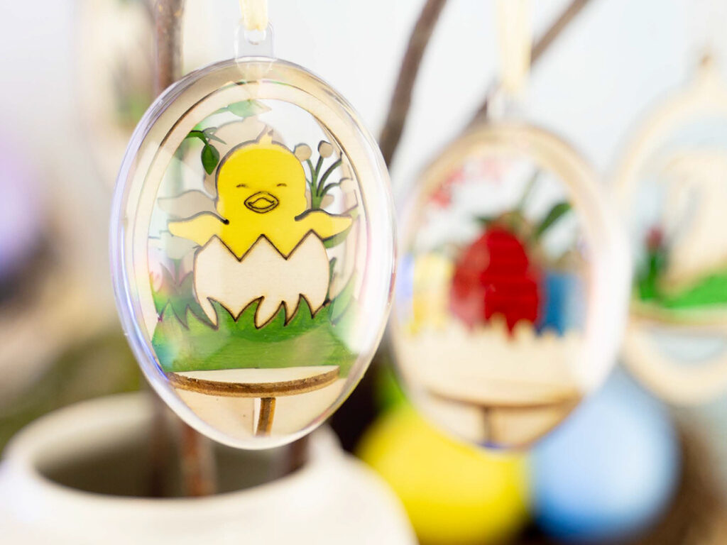 Gul kyllingfigur i egg av klar plast med utskåret tredekor, hengt på kvist.