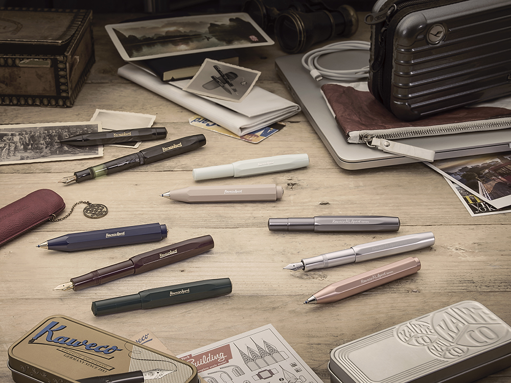Flere Kaweco-penner og tilbehør arrangert på trebord med postkort, etui og elektronikk